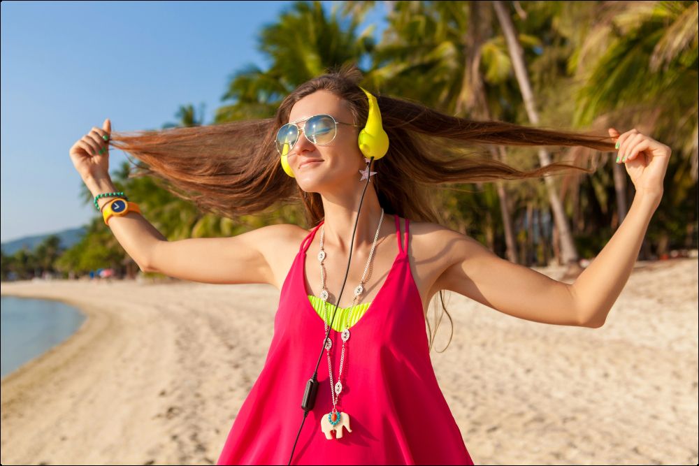 Foto de una mujer con cascos escuchando música en la playa que representa la idea de cómo cuidar el pelo en verano