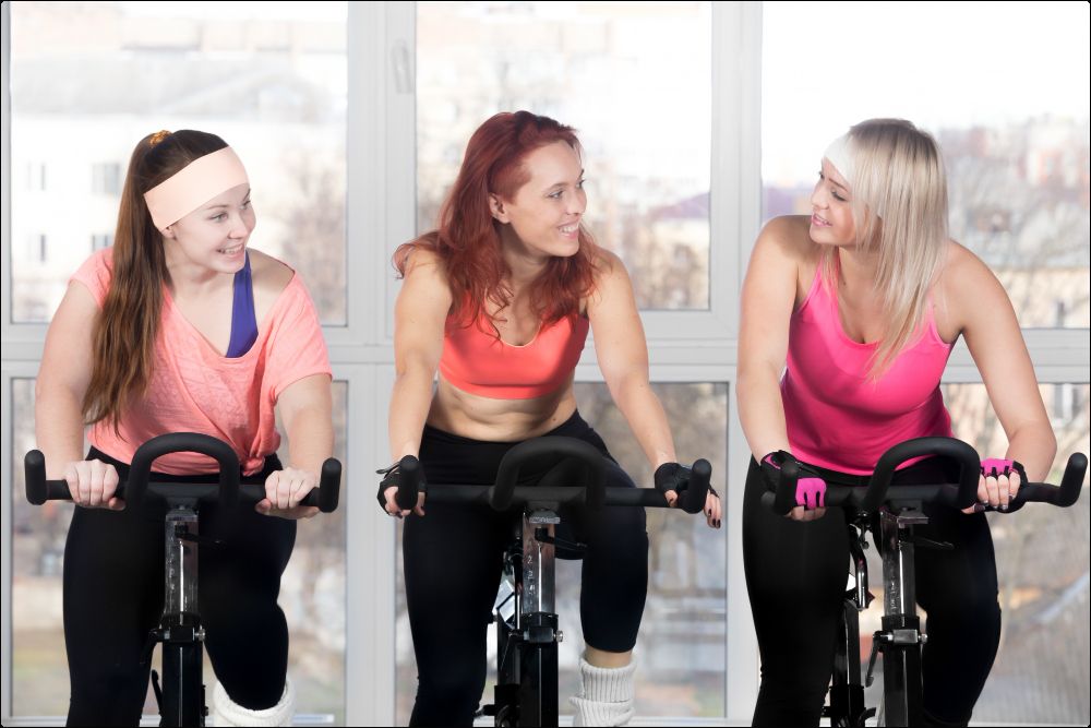 Foto de 3 mujeres sobre una bicicleta estática disfrutando de los beneficios psicológicos de hacer deporte